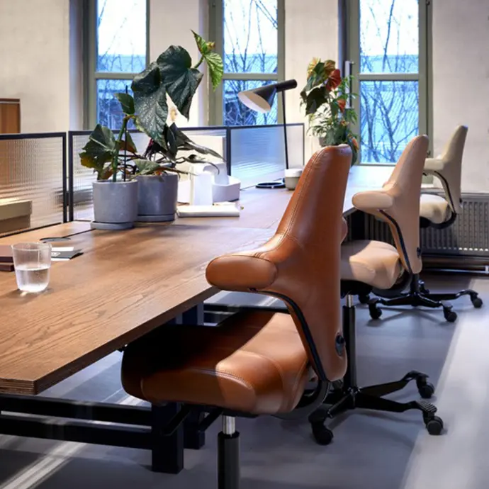 HÅG Capisco 8106 in cognacbruin leder — luxueuze uitvoering voor een professionele werkomgeving HÅG Capisco 8106 in cognacbruin leder aan houten vergadertafel in industrieel kantoor