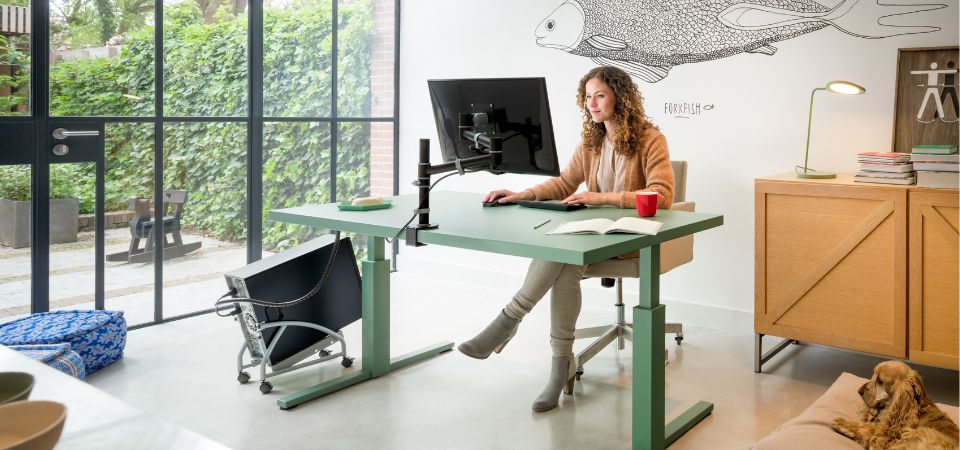 Moderne thuiswerkplek met een groene zit-sta bureau, ergonomische monitorarm en een kabelmanagementsysteem voor een opgeruimd bureau.