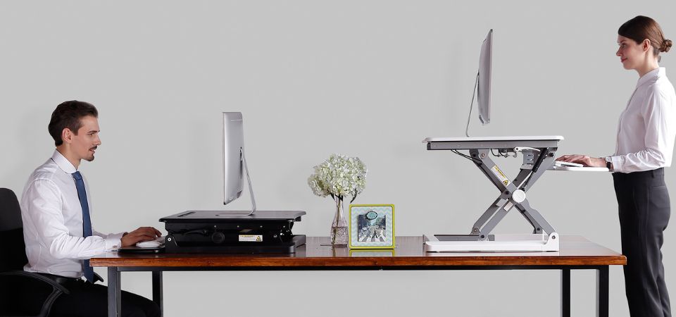Twee mensen werken met de Adjustable Sit-Stand Desk Riser 2: links zittend en rechts staand, wat het zit-sta principe illustreert.